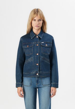 Woman with blonde hair wearing a dark blue denim jacket with copper buttons and light blue denim jeans, standing against a white background.