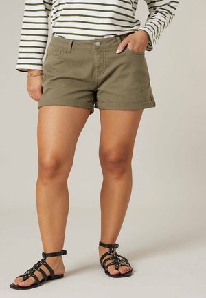 Femme portant un short vert olive à ourlet retourné, un haut à manches longues rayé et des sandales noires à bride à la cheville, debout avec une main dans la poche.