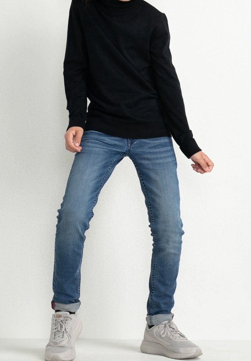 Personne portant un jean skinny bleu roulé, des baskets blanches et un haut noir à manches longues, debout contre un mur blanc uni.