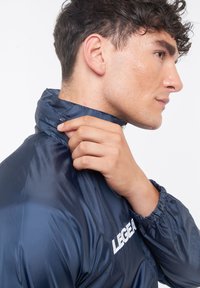 Giacca leggera navy con colletto alto, polsini elasticizzati e logo bianco prominente. Texture liscia e lucida, con zip frontale.