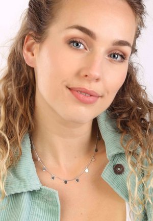 Zilveren ketting met ronde hangers, gedragen door een model met krullend haar, gecombineerd met een lichtgroen jasje van koord.
