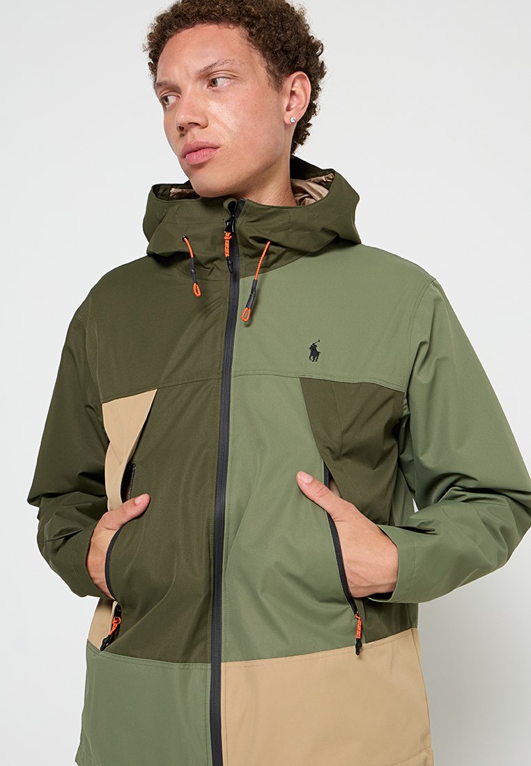 Giacca leggera con cappuccio in verde e beige. Caratterizzata da tasche con zip, cordini elasticizzati e un logo. Texture liscia e design a blocchi di colore.