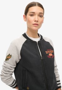 Veste bomber noire et grise avec fermeture éclair à l'avant, poignets côtelés et logo brodé sur la poitrine et la manche. Texture de tissu douce.
