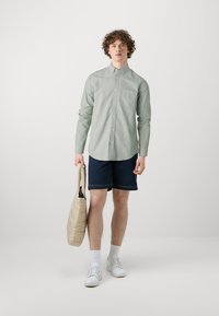 Lichtgroen button-up shirt, marineblauwe shorts, witte sneakers en een gewatteerde beige tote bag. Glad materiaal, casual fit en eenvoudig ontwerp.