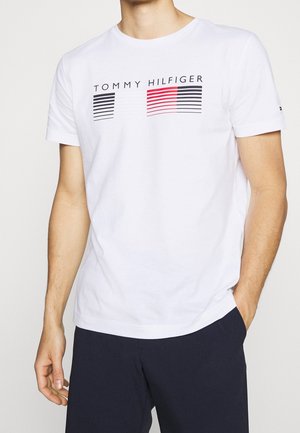 Weißes T-Shirt mit kurzen Ärmeln mit dem Schriftzug „Tommy Hilfiger“ und einem schwarz-weiß-roten gestreiften Muster, getragen von einer Person mit den Händen an den Seiten.