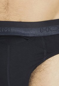 Nahaufnahme von schwarzen Herrenunterhosen mit grauem Bund am unteren Torso und oberen Oberschenkel einer Person.