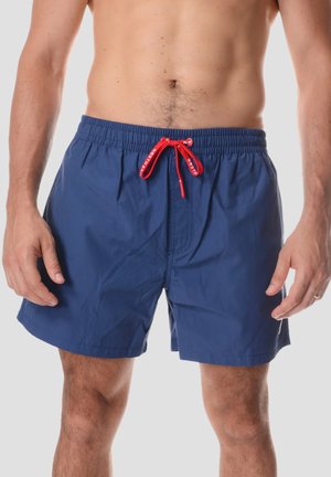 Uomo che indossa pantaloncini da bagno blu con coulisse rossa, in piedi con le mani rilassate ai lati contro uno sfondo semplice.