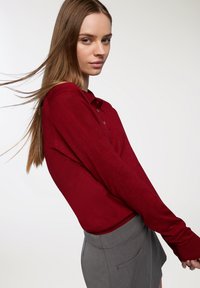 Dzianinowy czerwony golf z długim rękawem i kołnierzykiem, z trzema guzikami. Sweter ma gładką fakturę i luźny krój, połączony z szarym spódnicą.