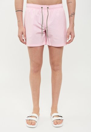 Rosa Badehose mit elastischem Bund, verstellbarem Kordelzug und kontrastierenden grünen Akzenten. Glattes, leichtes Material. Weiße Sandalen mit Riemen.