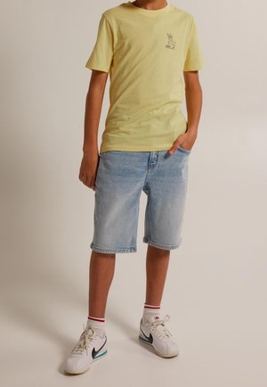 Žluté bavlněné tričko s malou grafikou na hrudi, spárované se světle modrými džíny a bílými teniskami s černými akcenty.