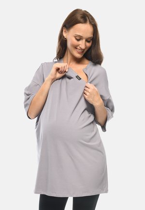Pregnant woman smiling, wearing loose gray top, pulling open neckline to reveal hidden nursing access.