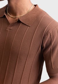 Braun strukturiertes Polohemd mit Kragen, kurzen Ärmeln und vertikalen Rippmustern. Eine goldene Kettenhalskette betont den Look.