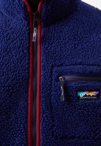 Fleecejakke i dyb marineblå med en tekstureret overflade. Har en maroon lynlås og en sidelomme med en sort lynlås, mærket "Manastash."