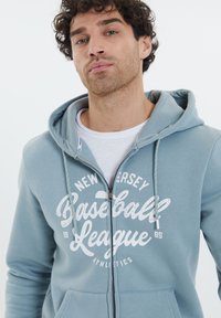 Helle blaue Zip-Hoodie aus weichem Material, mit einer Fronttasche, einer Kordelzugkapuze und einem weißen "Baseball League" Grafikdesign.