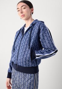 Marinblå zip-up hoodie med geometriskt mönster, ribbade ärmslut och nederkant. Har en huva med dragsko och en liten logotyp på bröstet.