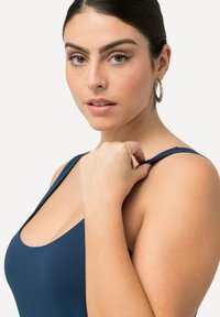 Námořnická modrá body s kulatým výstřihem a tenkými ramínky, vyrobená z hladkého, elastického materiálu. Modelka se dotýká ramínka rukou.