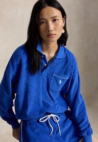 Joven mujer con cabello largo y oscuro que lleva una camisa polo de manga larga azul y pantalones a juego con cordón, de pie frente a un fondo neutro.