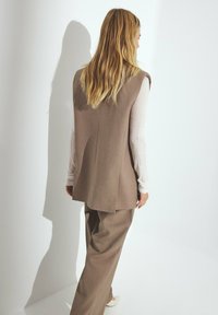 Lichtbruine op maat gemaakte vest met een gelaagd ontwerp, gestyled over een langslavige beige top en een bijpassende broek; gladde stoftextuur.