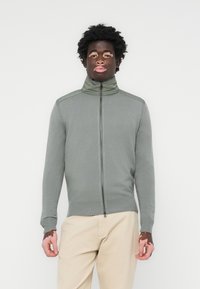 Zip-up sweater in muted green, made of ribbed fabric. Features a high collar, two-tone shoulder accents, and fitted cuffs. Minimal design.