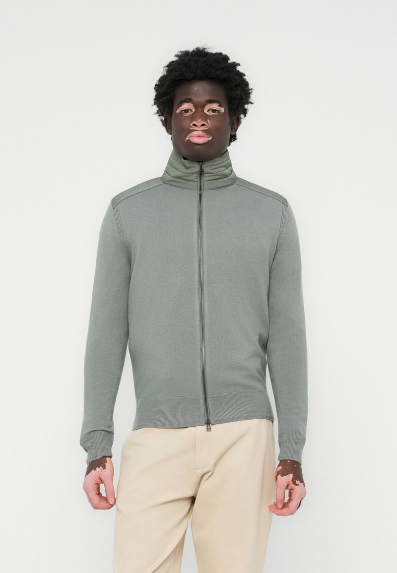 Zip-up sweater in muted green, made of ribbed fabric. Features a high collar, two-tone shoulder accents, and fitted cuffs. Minimal design.
