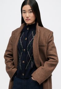 Cappotto intrecciato marrone con colletto a scollo a V sopra un cardigan a motivo argyle blu navy. Chiusura con bottoni e jeans di denim scuro completano il look.
