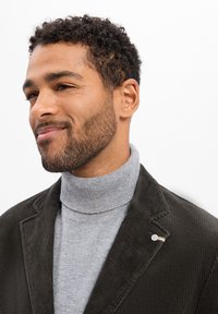 Dunkelgrüner Cordblazer über einem hellgrauen Rollkragenpullover. Der Blazer hat einen dekorativen Knopf mit Streifen und zeigt einen strukturierten Stoff.