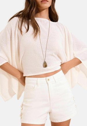 Femeie care poartă un top larg tricotat alb, pantaloni scurți albi cu talie înaltă și șiret lateral, și un colier lung cu pandantiv, mâinile sprijinite pe șolduri.