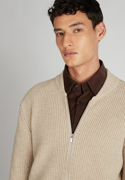 Beige geribbelde cardigan met een ritssluiting en brede kraag, laag over een bruin overhemd met knopen. Soepele textuur, casual ontwerp.