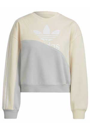 Woman wearing cream and gray Adidas sweatshirt with trefoil logo and matching cream pants, hands in pockets, cropped view.