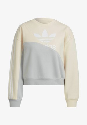 Femme portant un sweat-shirt Adidas crème et gris avec logo trefoil et pantalon crème assorti, mains dans les poches, vue recadrée.