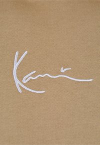 Tan stoff med et fremtredende, glatt hvitt brodert signaturdesign. Teksturen er strikket med et fint, ensartet mønster.