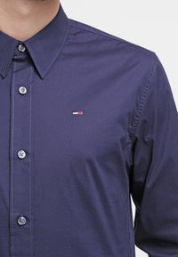 Camicia abbottonata blu navy con colletto classico, cuciture chiare e un piccolo logo ricamato sul petto. Tessuto di cotone liscio.