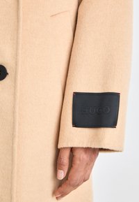 Manica di un cappotto di lana beige con una toppa rettangolare nera con l'etichetta "HUGO" e una mano visibile sotto la toppa.