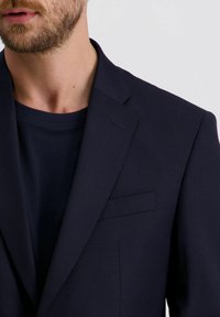 Behaarde man die een marineblauwe blazer over een bijpassend marineblauw shirt met ronde hals draagt, weergegeven van de nek tot de schouder.