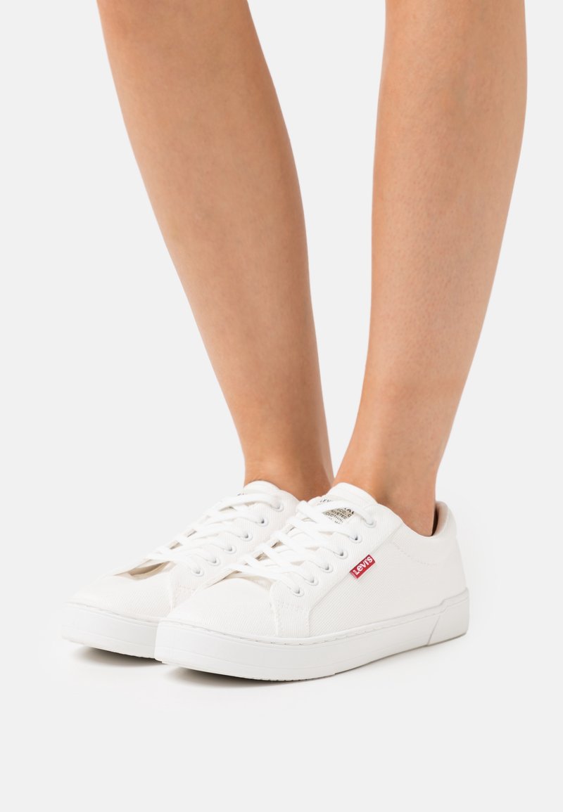 Zapatillas blancas de lona con diseño de cordones, que presentan un pequeño logotipo rojo de Levi's y una suela plana de goma. Superficie texturizada y puntera redondeada.