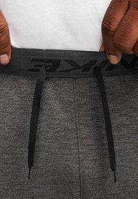 Mains à la peau foncée ajustant le cordon noir de la ceinture d'un pantalon de survêtement gris sur un t-shirt blanc.
