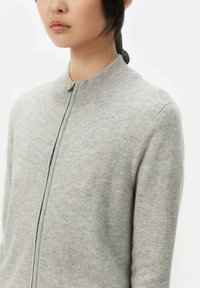 Grå stickad tröja med hög krage och främre dragkedja, med en slät yta och figurnära design. Minimalistisk stil, inga synliga mönster.