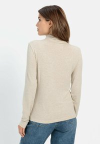 Beige Langarmshirt mit weicher Textur und tailliertem Design. Hat einen hohen Ausschnitt und wird mit blauen Jeans kombiniert.