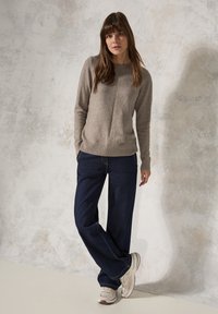 Hellbrauner Pullover mit gerippten Bündchen und Saum, kombiniert mit dunkelblauen weit geschnittenen Jeans und beigen Sneakern, vor einem strukturierten hellen Hintergrund.