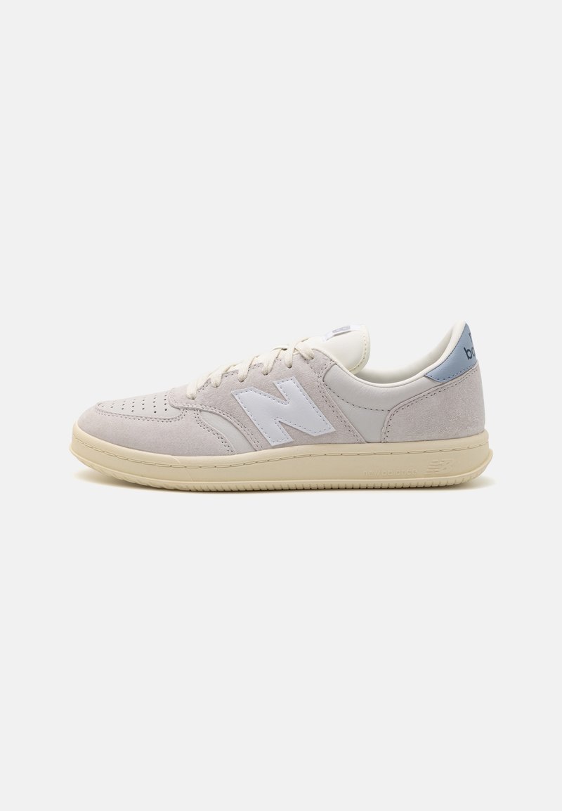 Baskets New Balance beige clair avec logo blanc « N », languette de talon bleu clair, bout perforé et semelle couleur crème sur fond blanc.