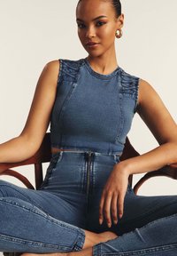 Denim crop-top, jossa on olkaindetallia, yhdistettynä korkeavyötäröisiin farkkuihin, joissa on etuvetoketju, keskisinisellä pesulla.