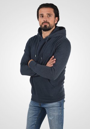 Tamno plavi dukserica od mekog materijala s kenguru džepom, kapuljačom s vezicama i rebrastim manžetama, u kombinaciji s iskrzanim svijetlim trapericama.