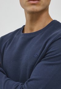 Navy crew neck sweatshirt with ribbed collar, soft texture, and a small embroidered logo on the chest. Minimalist design.