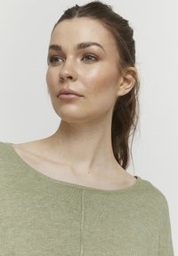 Svetle zelený, strukturovaný pletený top s širokým, kulatým výstřihem. Obsahuje jednoduché linie a minimální detaily stehování, což zdůrazňuje pohodlný design.