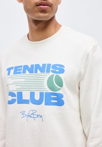Άνω κορμός ενός ατόμου που φοράει λευκό φούτερ με μπλε κείμενο "TENNIS CLUB" και πράσινο γραφικό με μπάλα του τένις μπροστά.