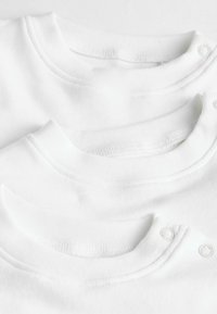 Tre hvide bomulds t-shirts ligger stablet, med runde kraver og trykknapsdetaljer på en af dem, samt en glat, strækbar tekstur.