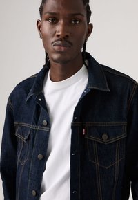 Ciemnoniebieska kurtka jeansowa z kontrastowym żółtym przeszyciem, dwoma kieszeniami na piersi, zatrzaskami oraz białą koszulką z okrągłym dekoltem pod spodem.