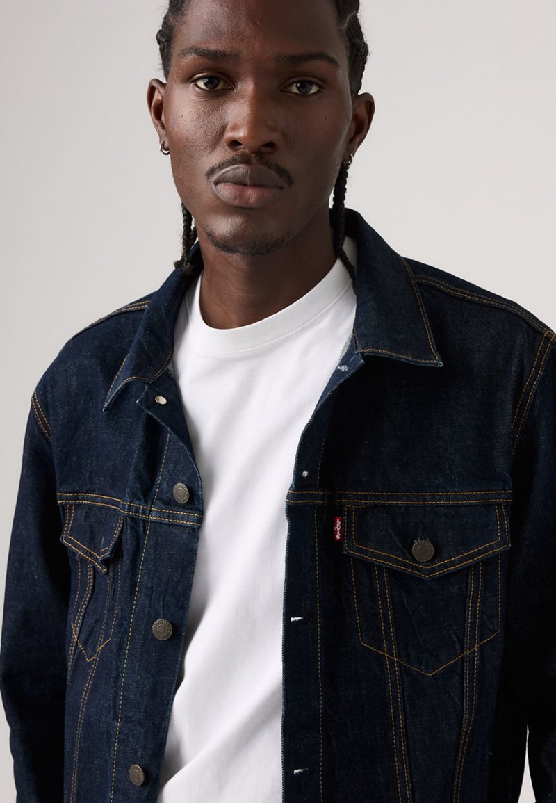 Dark blue denim jacket featuring contrast yellow stitching, two chest pockets, snap buttons, and a white crew neck t-shirt underneath.