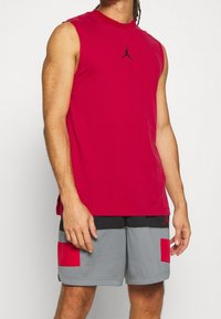 Rode mouwloze sportshirt met een zwart logo in het midden van de borst, gecombineerd met grijze shorts met rode accenten en zwarte details.