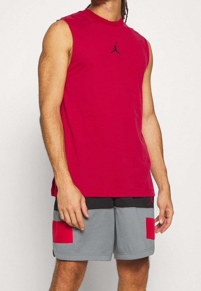 Rode mouwloze sportshirt met een zwart logo in het midden van de borst, gecombineerd met grijze shorts met rode accenten en zwarte details.